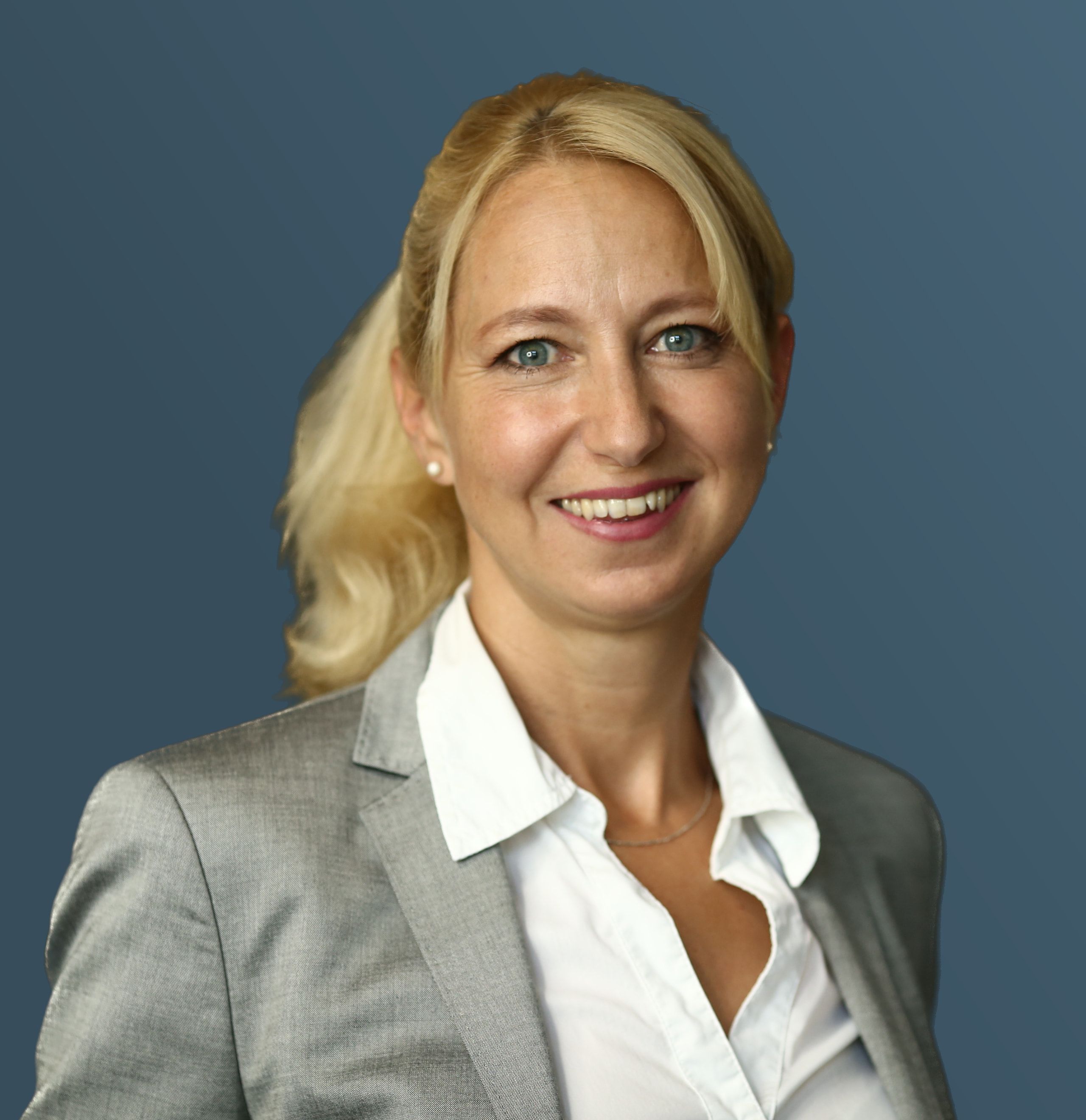 Daniela Meingaßner, lächelnd, im grauen Blazer vor blauem Hintergrund